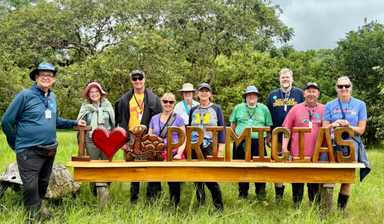 A group of travelers stand in front of a wooden sign reading "I heart primicitas"