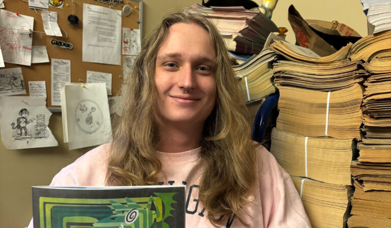 Luke Homans, with long blond hair and a light pink shirt, poses with a green issue of the Gargoyle.