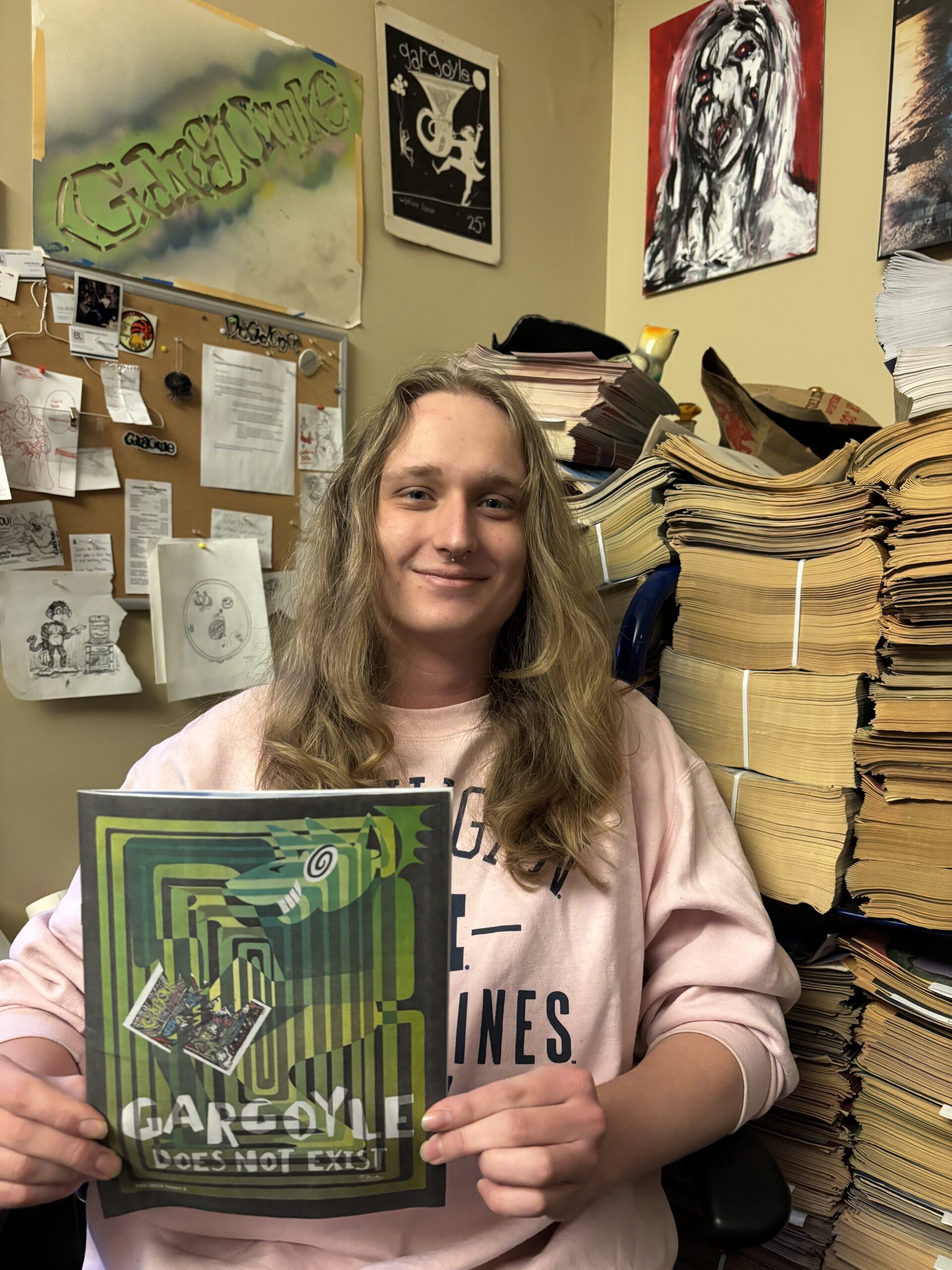 Luke Homans, with long blond hair and a light pink long sleeve shirt, poses with an issue of the Gargoyle with stacks of old paper behind him. 