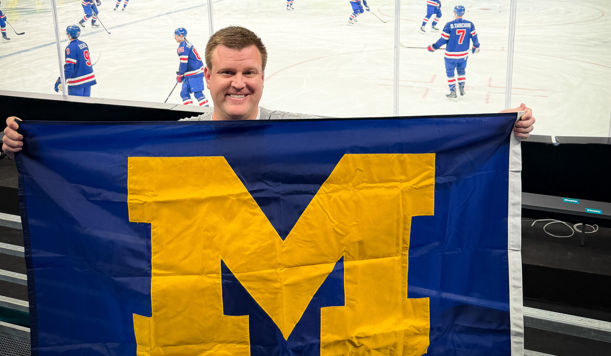 Austin Chapman poses with a Block M flag in front of him and the US men's hockey team warming up behind him.