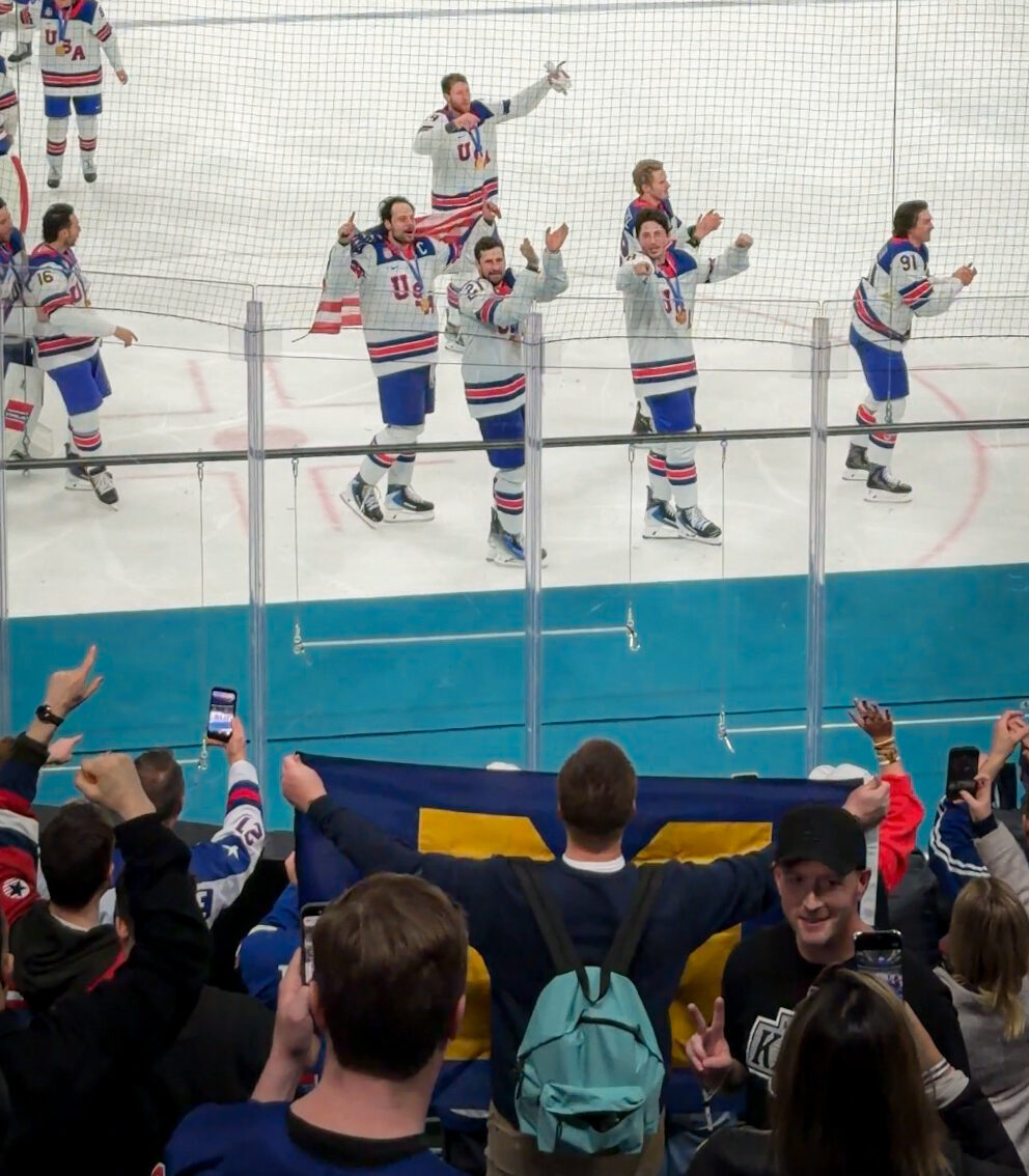 Chapman, holding the Block M flag, faces away from the camera in an ice rink crowd. Some of the players on the ice point towards him.