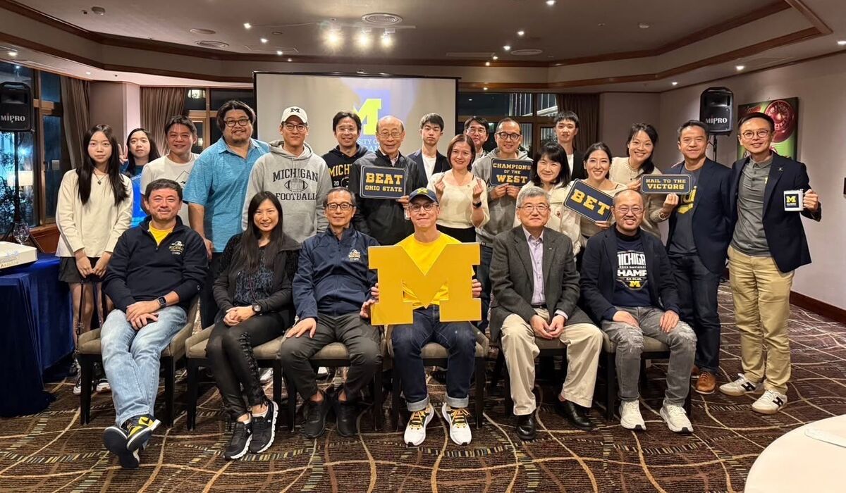 A group from the U-M Club of Taiwan pose for a picture. The person in the middle is holding up a large block M as they sit inside a hotel room conference center.