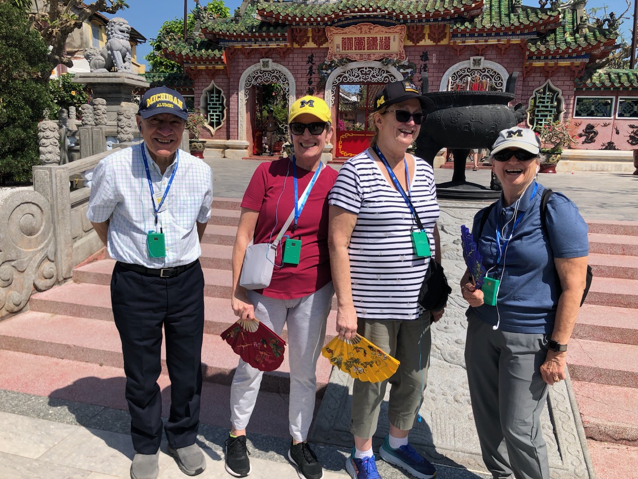 Four people (three women and a man on the right, pose in Vietnam. They are all wearing U-M hats and green badges on lanyards. 