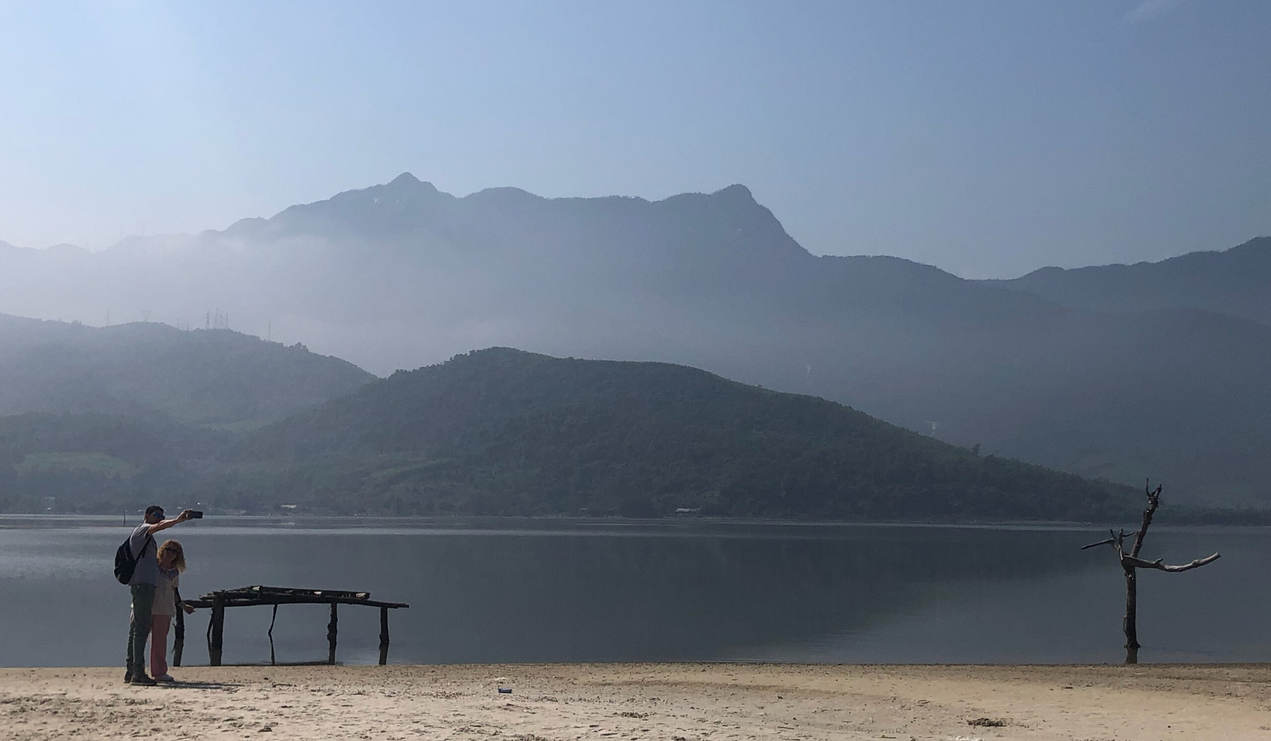 Foggy mountains in the distance with sand in the foreground and water in between. Two people stand on the sand taking a photo.