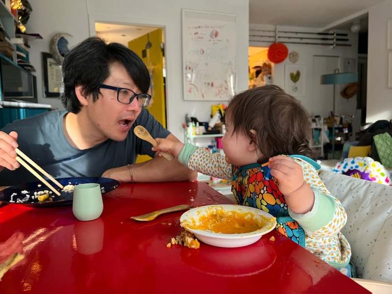 John Wang sits at a red table with his daughter who is reaching toward him with a wooden spoon. They are eating together. 