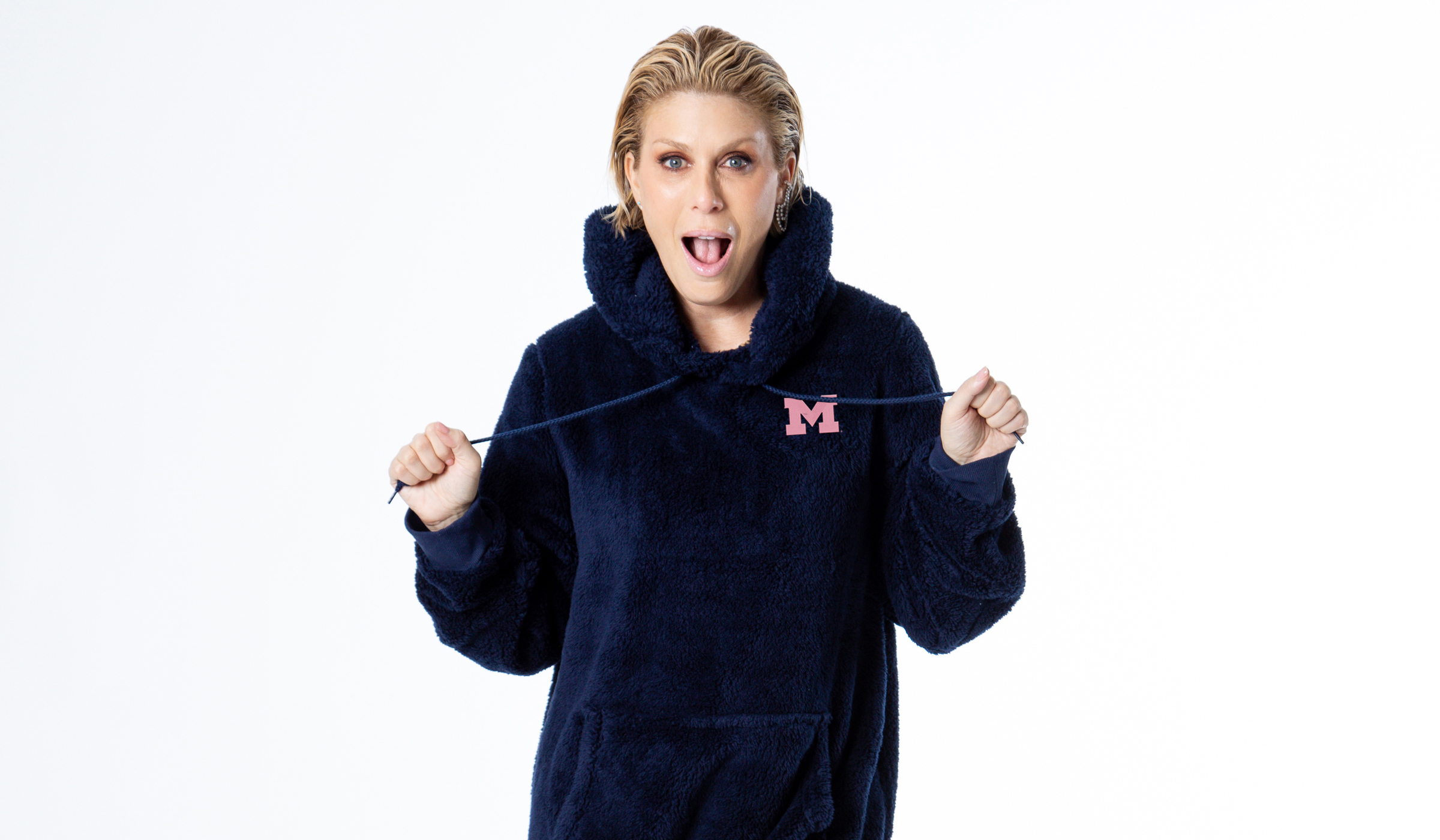 A woman stands with a surprised look on her face in an oversized fleece jacket that has a Michigan block M on it that is pink.