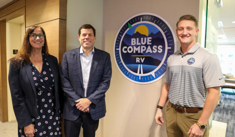 Jon Ferrando (center) poses with Shahnaz Broucek (left) and Jared Davis (right). The Blue Compass RV logo is in the middle.