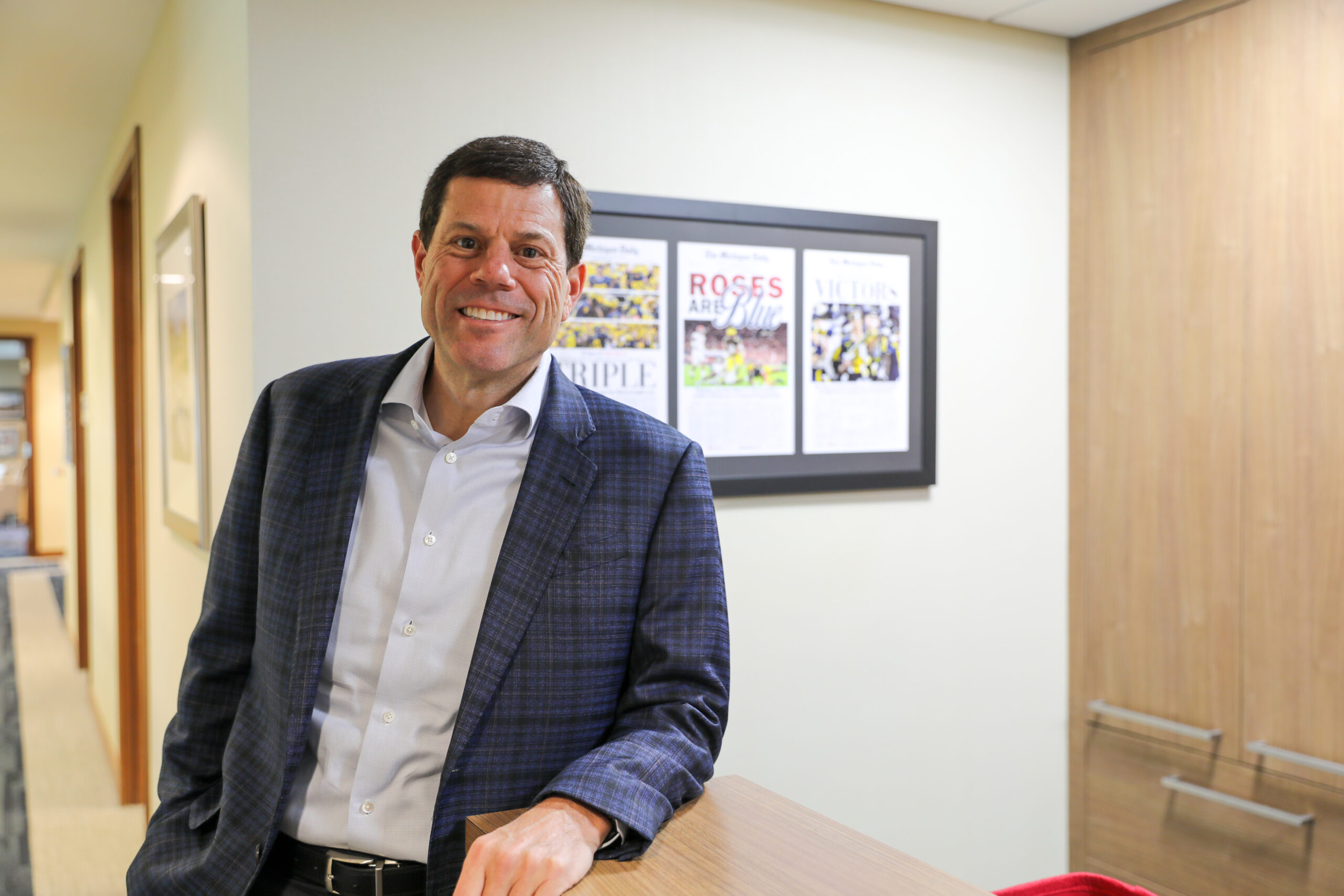 Jon Ferrando, wearing a light blue button-down and blue blazer, leans on a desk with his left arm.