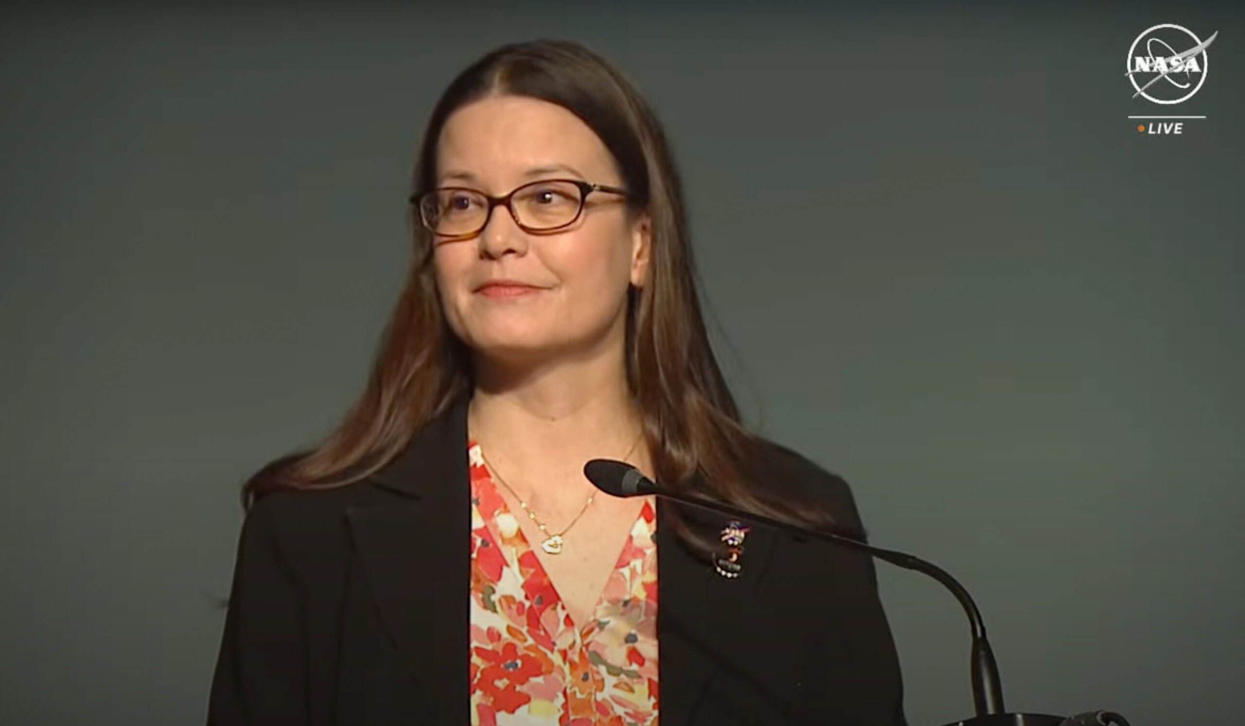 Kelly Korreck, wearing glasses, a flowery shirt, and a black blazer, speaks at a podium on a grey backdrop.