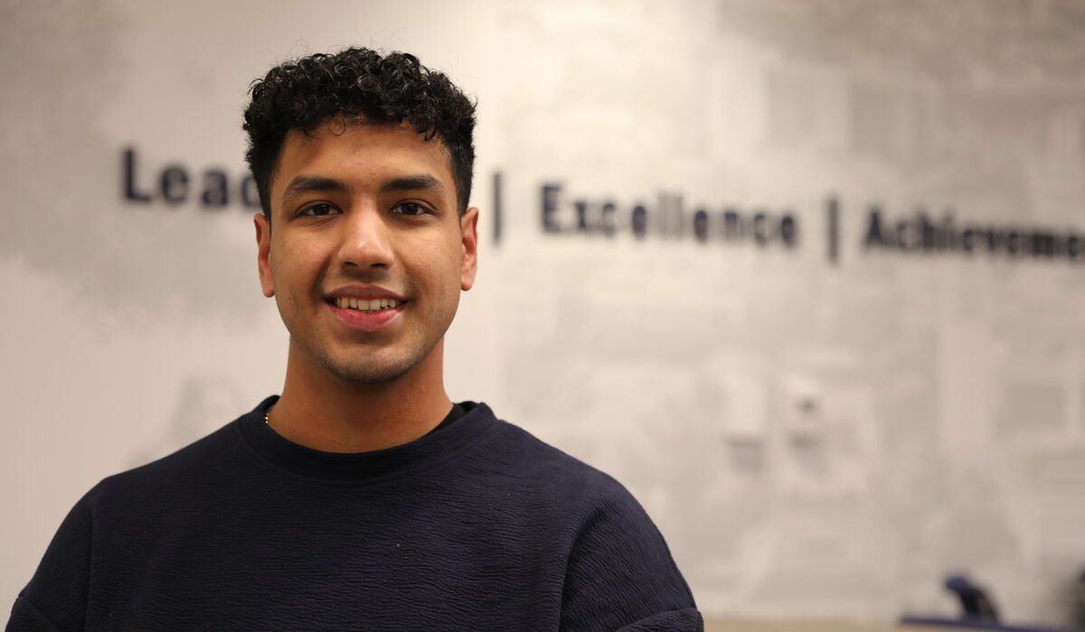 Photo portrait of Devon Krawczyk smiling toward the camera. Behind him is a wall featuring non-descript photographic elements and the words Leadership, Excellence, and Achievement (partially trailing out of the image to the right).