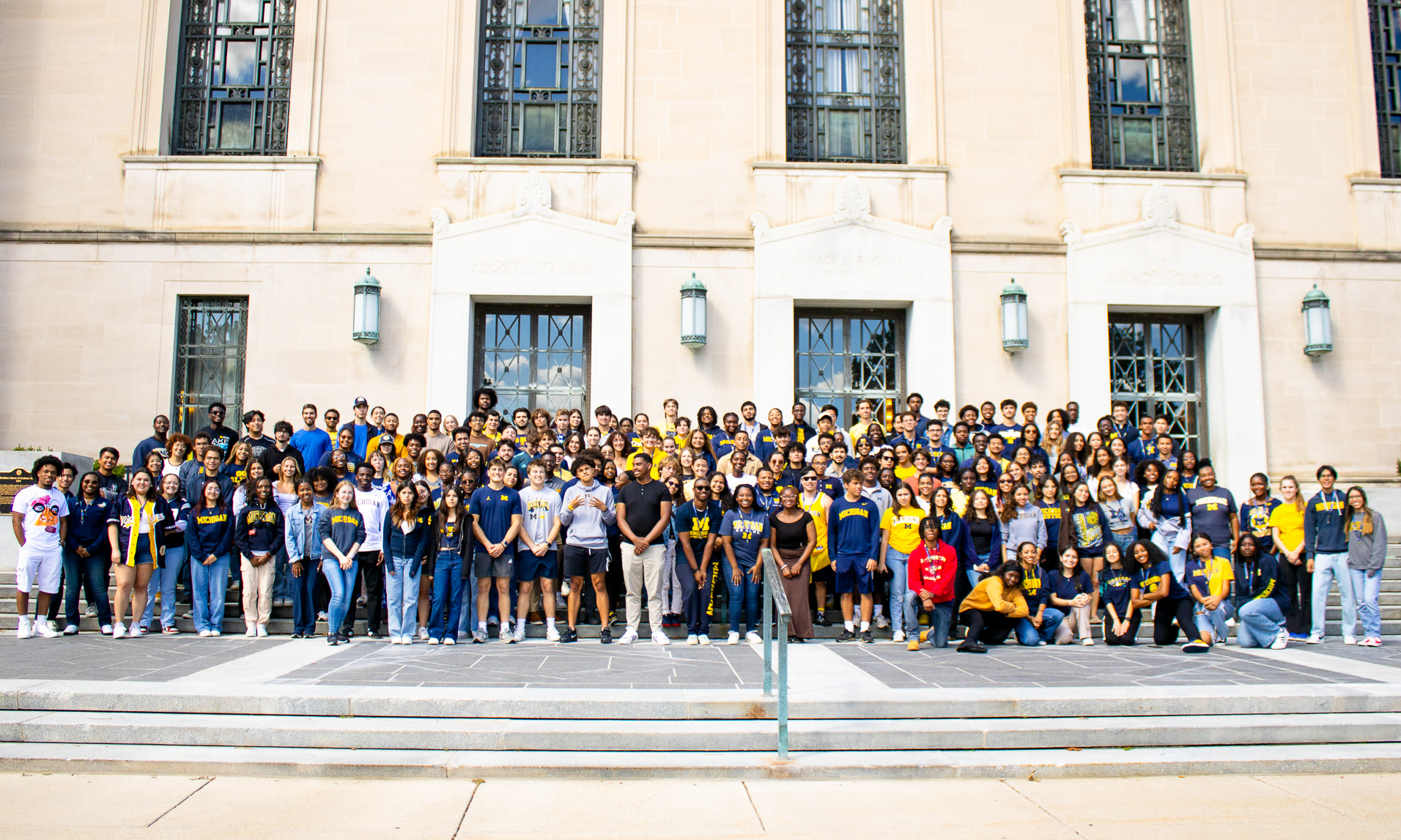 A large group of students stand in formation in front of the Rackham Graduate School at the University of Michigan.