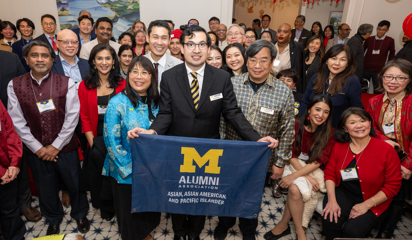 A large group of people pose for a photo. In the center, a man in a suit is holding a Michigan flag.