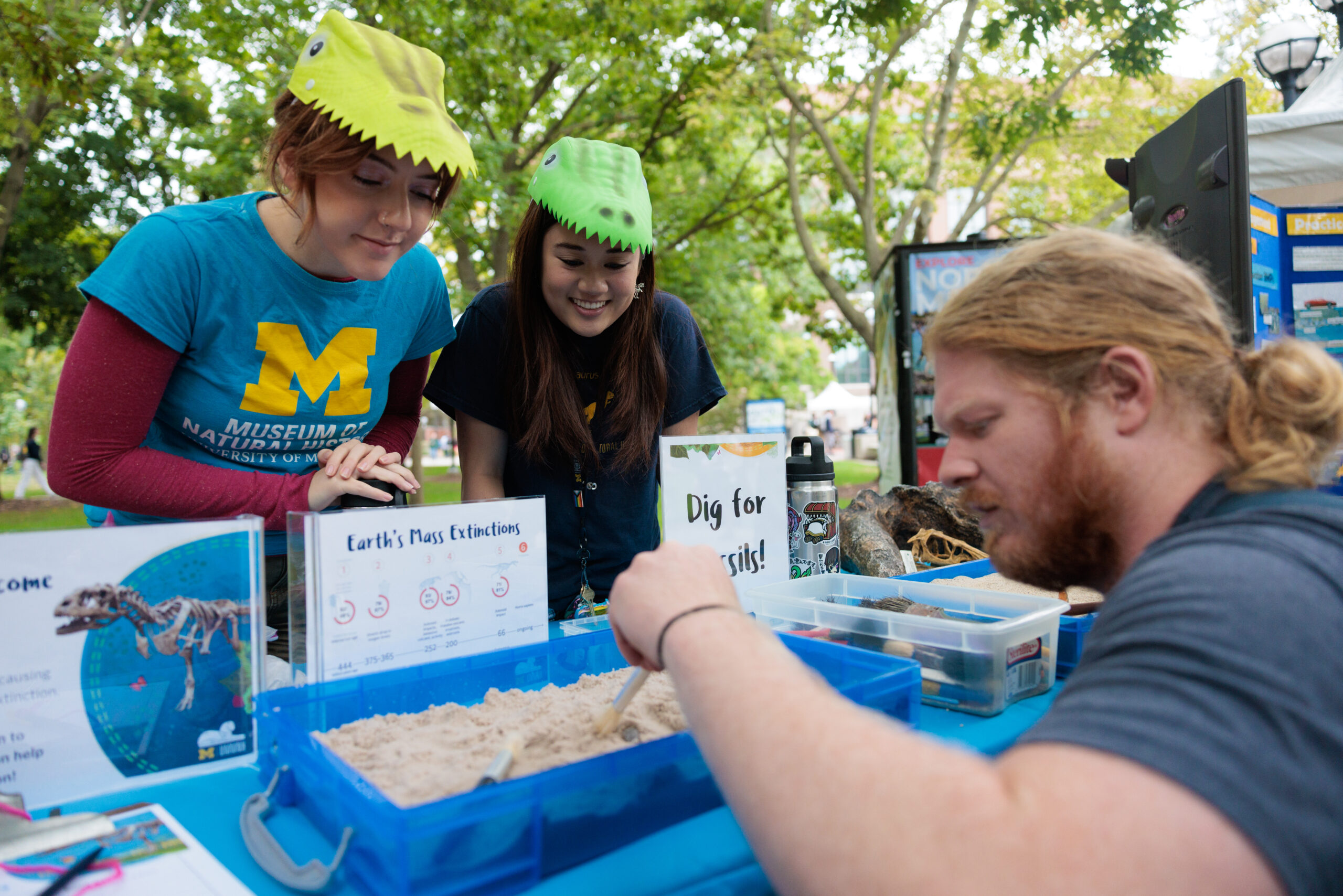 Volunteers from the University of Michigan Museum of Natural History wearing plastic dinosaur hats watch as a student uses tools to dig through a small container of sand looking for fossils at EarthFest 2024.
