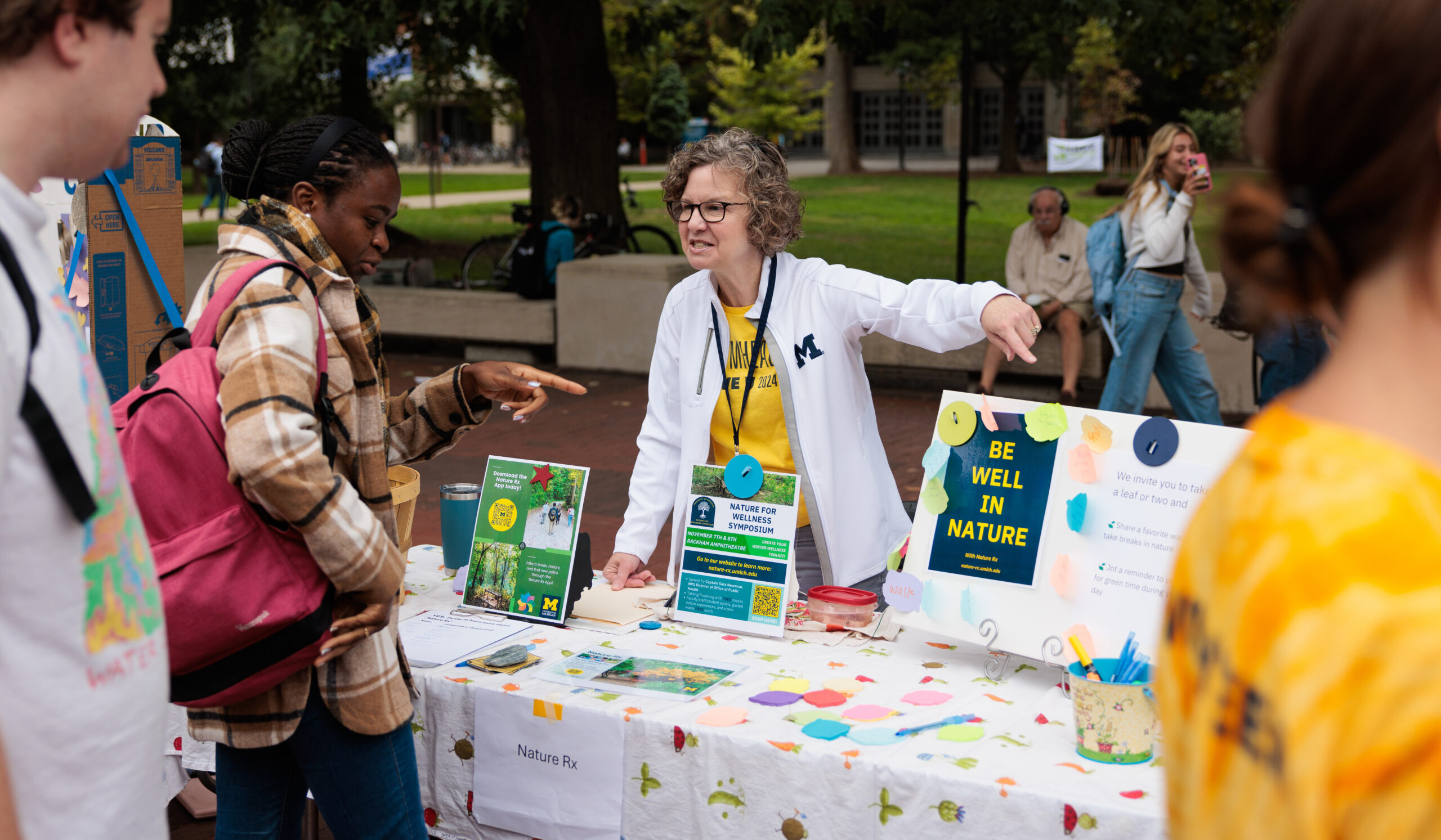 An employee from NatureRX points toward an info board reading “Be Well In Nature” while talking to students at EarthFest 2024.