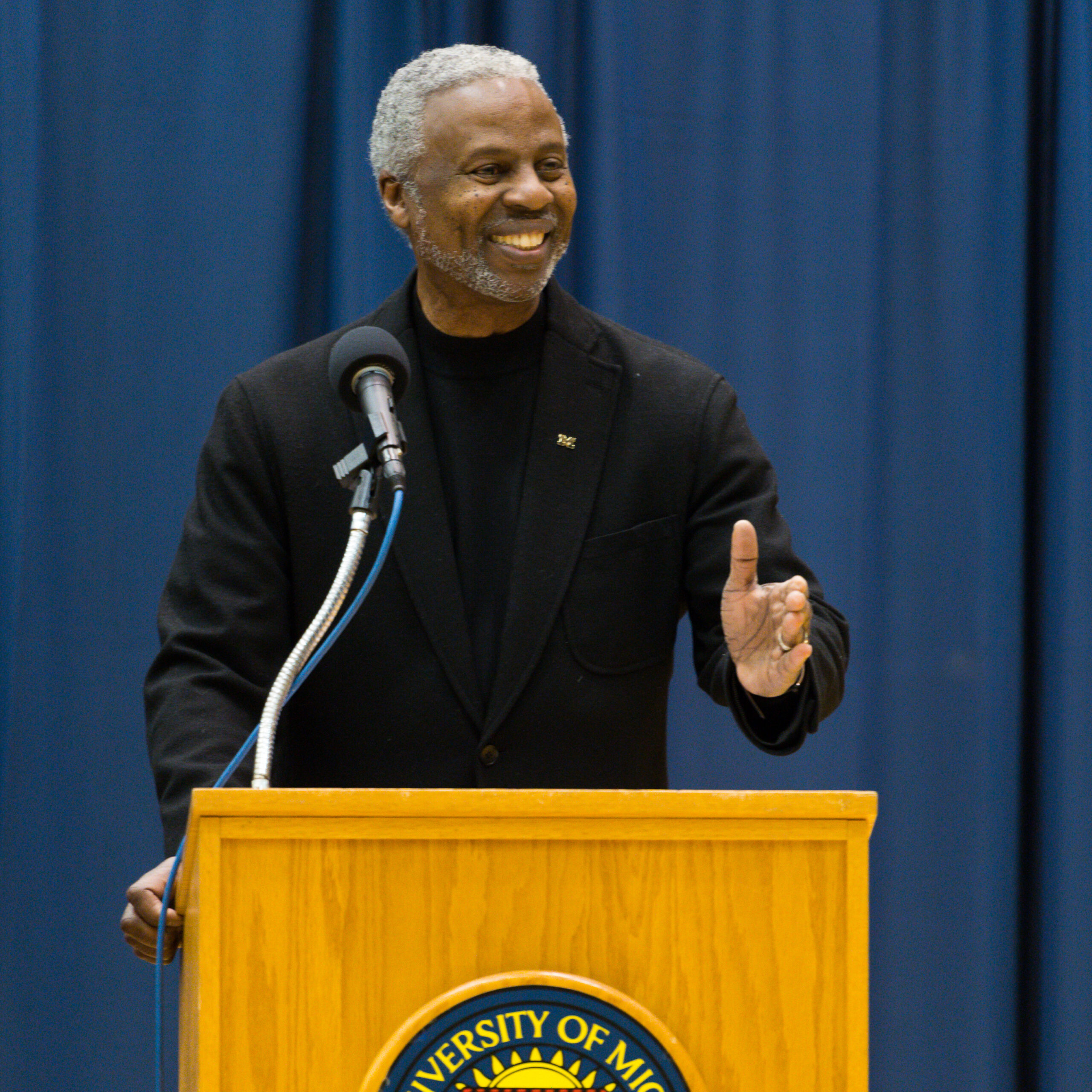 Greg Harden in all black standing at a podium with a blue background.