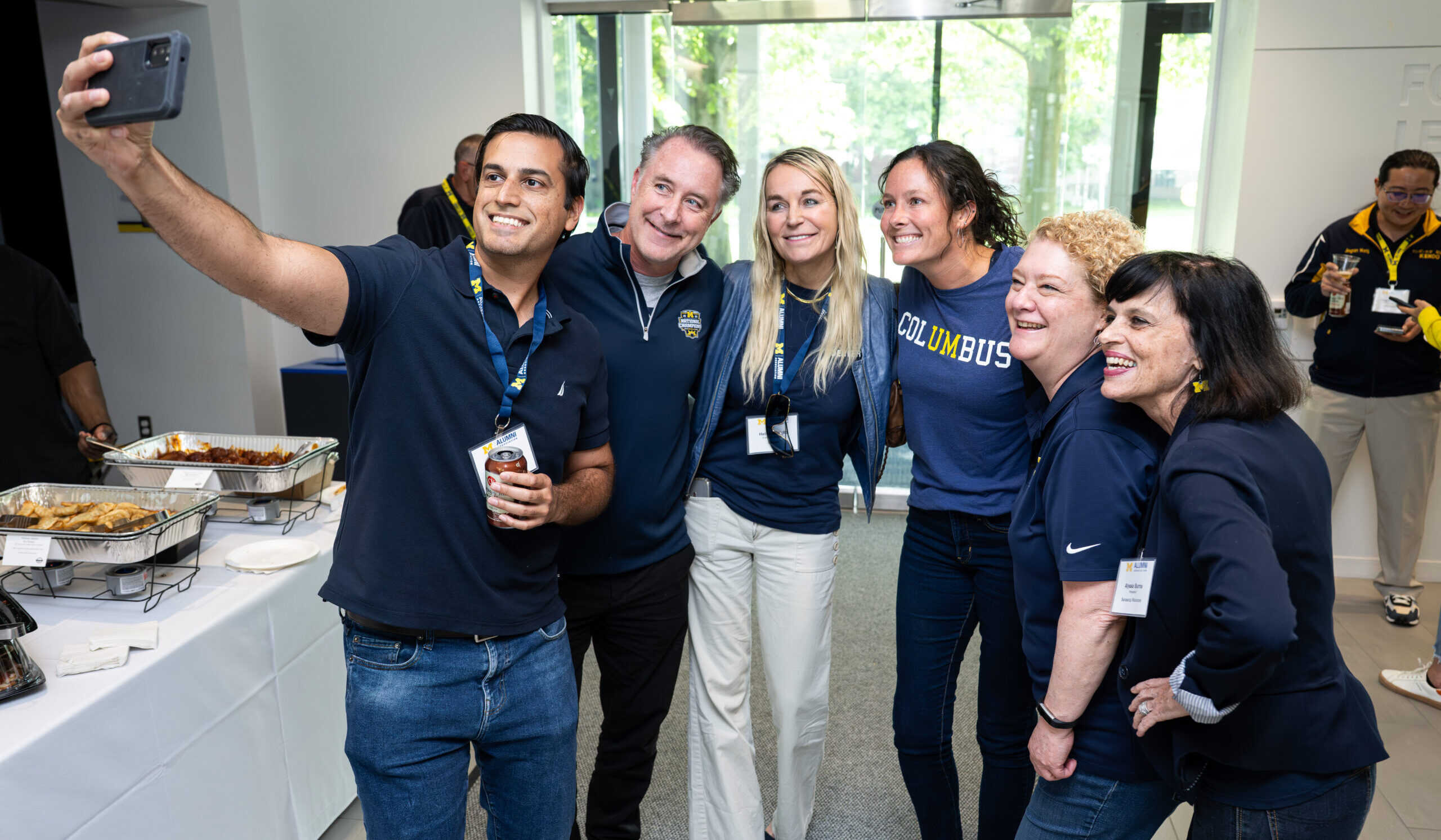 Alumni and staff all in blue shirts pose for a selfie, with the camera held by the person on the far left.