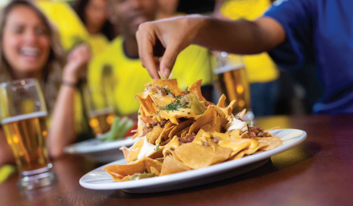 Image of a hand pulling a chip from a plate of nachos, people in yellow and blue in the background