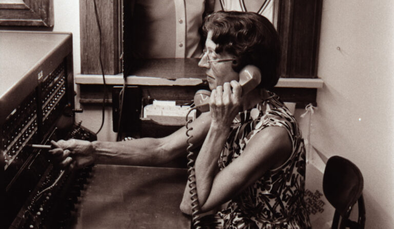 A black and white photo from 1975 showing a woman with short hair and a phone in her left hand making a phone connection on a switchboard.