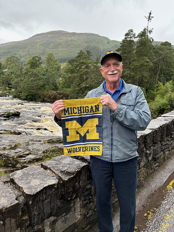 Mark Mazur, ’75, beams with Maize and Blue pride while posing at the Falls of Dochart in Scotland during an Alumni Travel trip in August 2024.