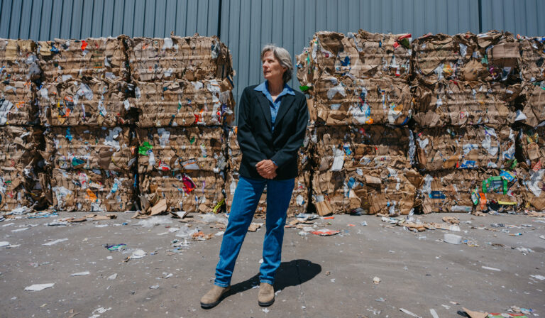 Zan Jones, in jeans and a dark blue blazer, stands with her hands together in front of compacted recycled materials.