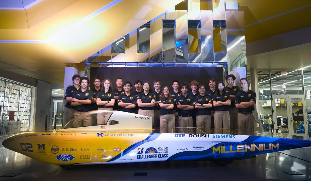 All 18 members of the solar car race crew stand behind Millennium with their arms crossed. Most of the vehicle is roughly knee high and covered in solar cells, with the top of the cockpit at hip height.