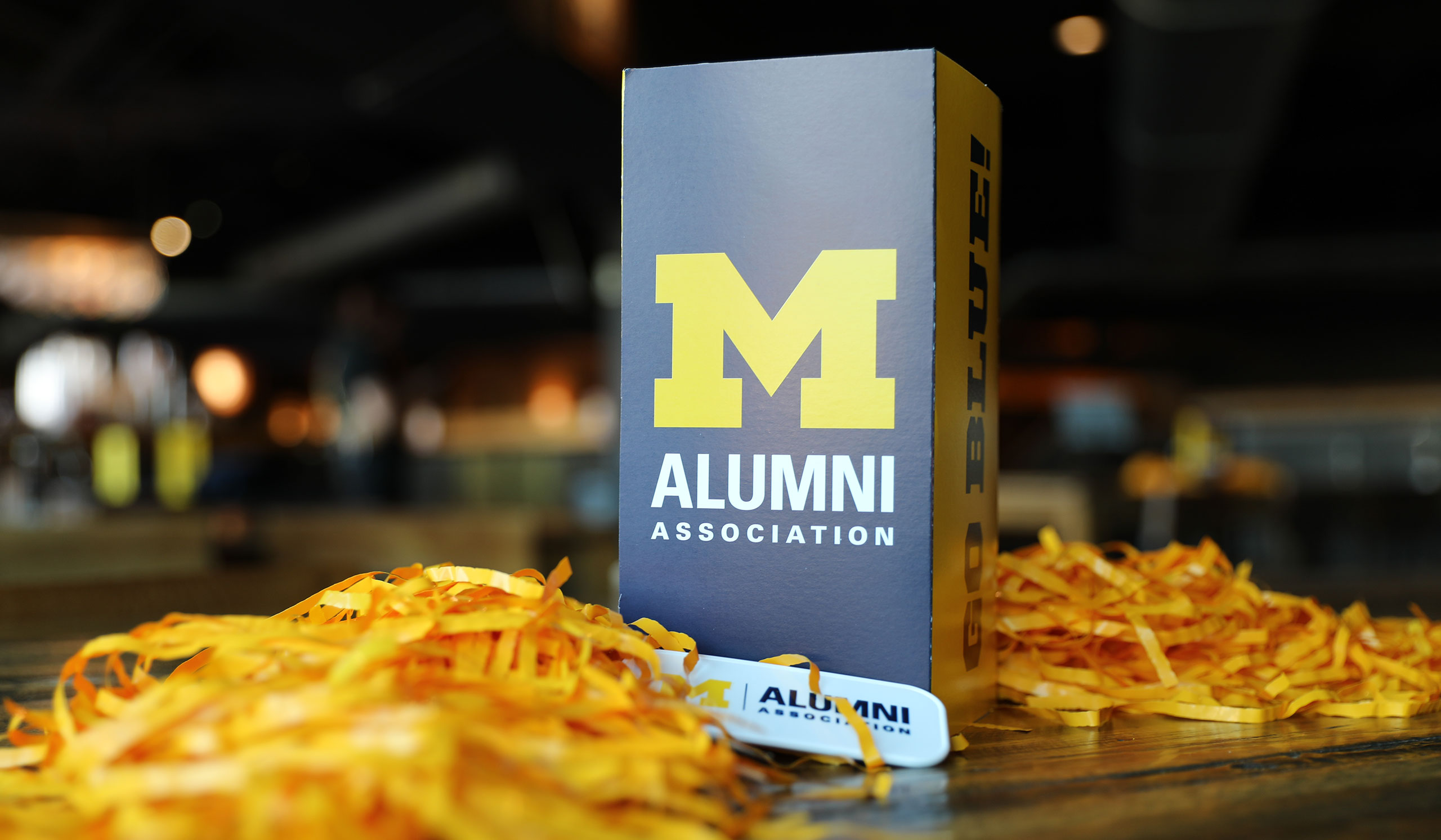 Michigan table decorations at a bar, including maize pompoms and a standing sign with the logo for the Alumni Association of the University of Michigan on one side and "GO BLUE" on another.