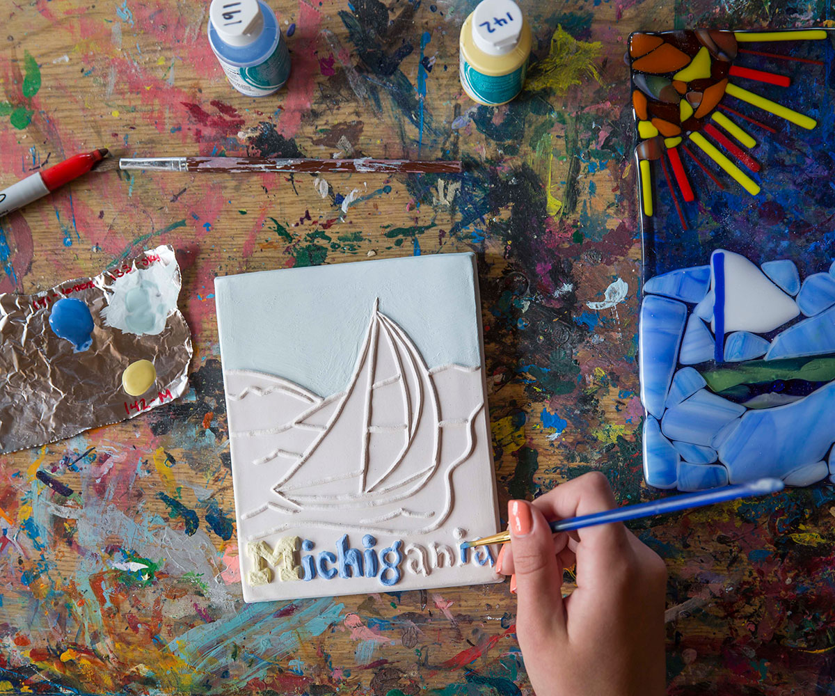 A person paints a ceramic tile of a sailboat and the word Michigania