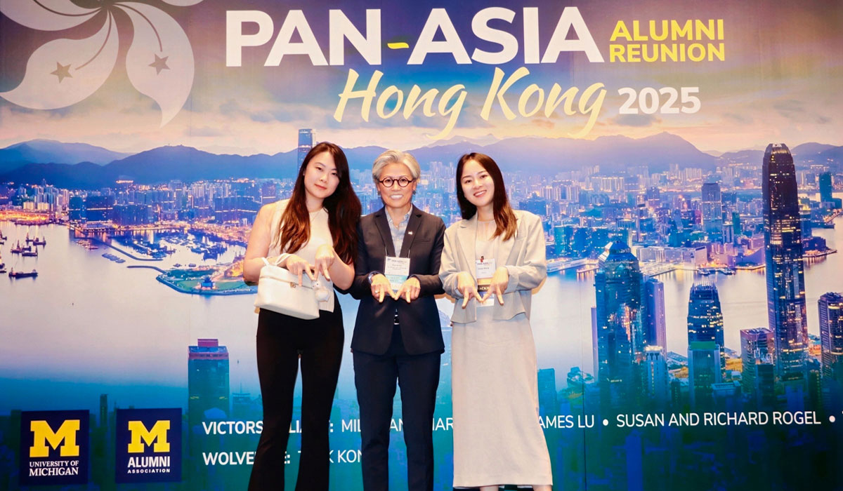 Three people pose for a photo in front of a backdrop at the 2025 Pan-Asia Alumni Reunion. They're all forming an "M" with their hands.