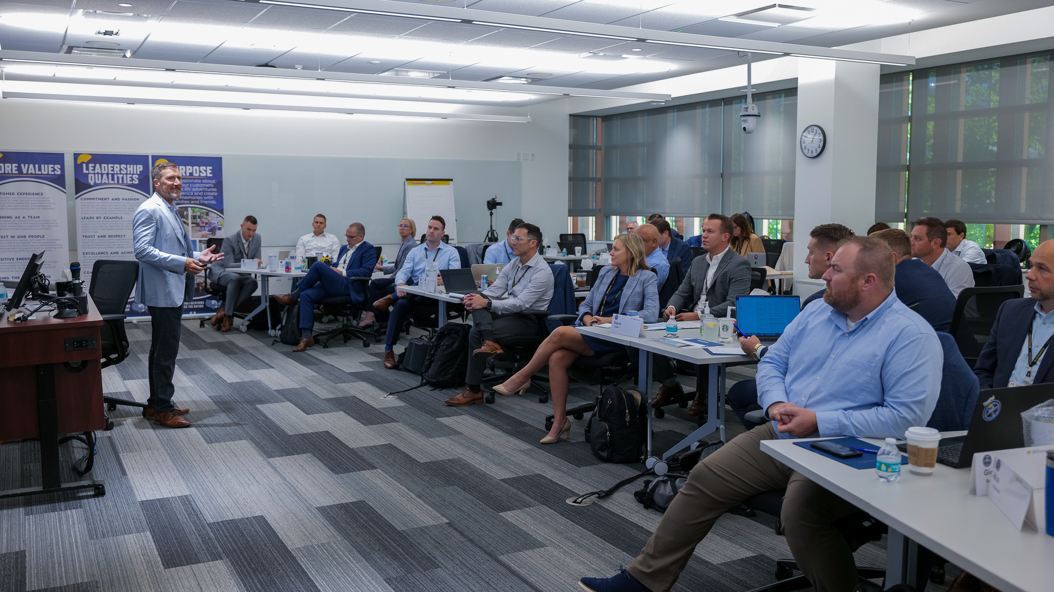 A room of business people sit on the right side with someone speaking to them on the left.