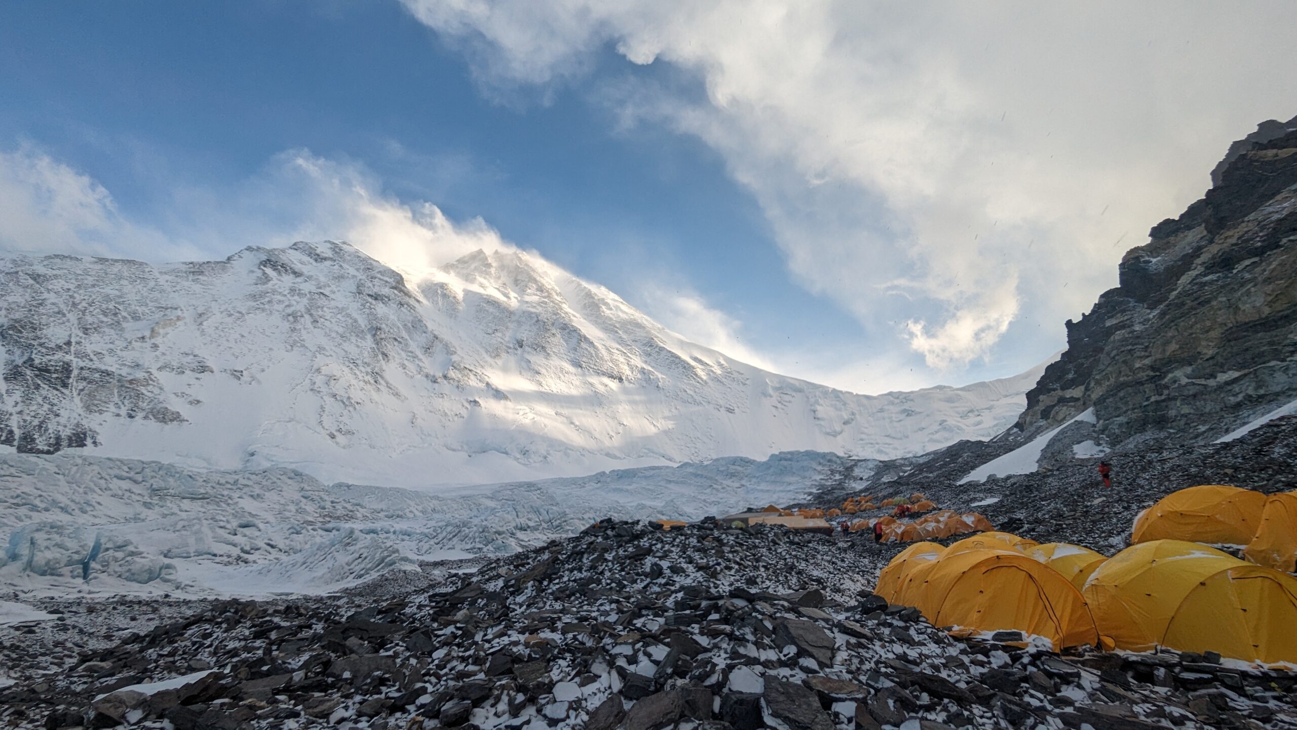 A wide image with a snowy Everest on the left and rocky terrain with yellow tents on the right.