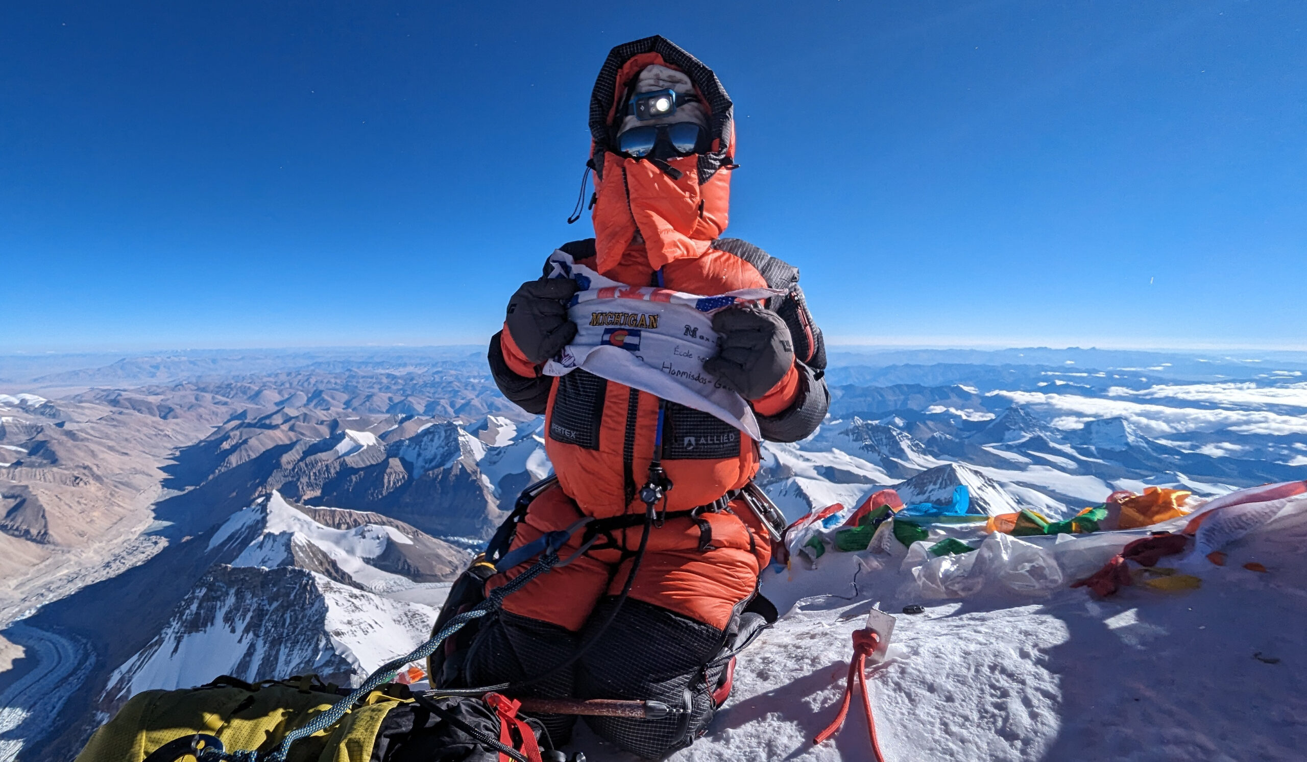 Myra Beaudoin Bertrand in an orange winter suit, kneels on top of Everest and holds a homemade flag.