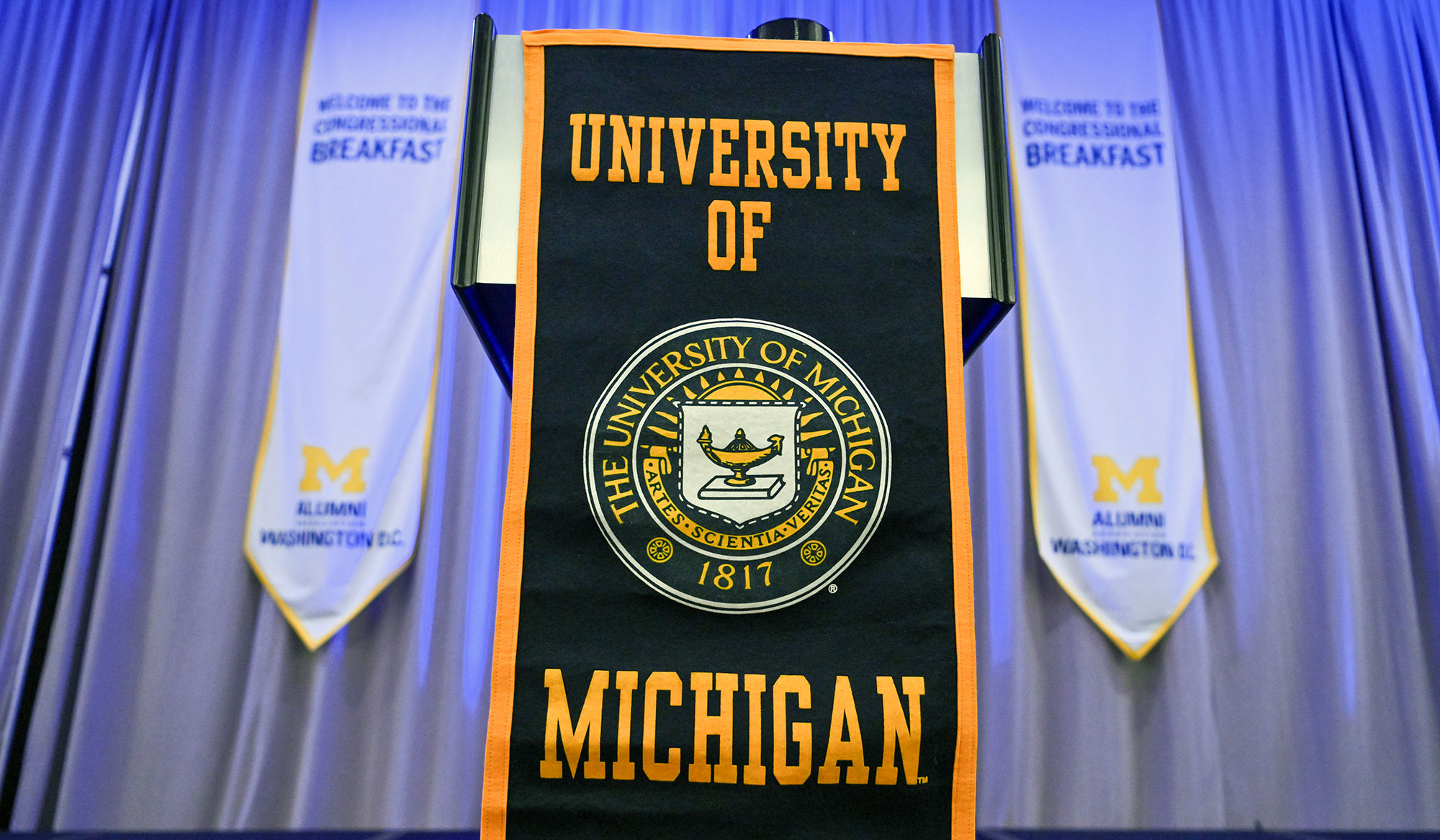 A podium draped with a University of Michigan banner stands between two tall banners reading “Welcome to the Congressional Breakfast,” with logos for the U-M Club of Washington D.C. below.