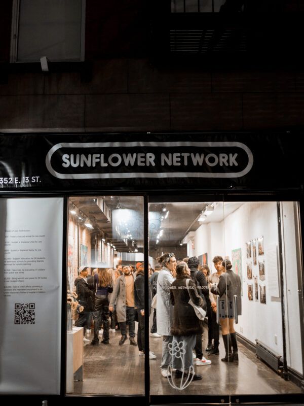 Night-time exterior photo looking into the “Sonya: A Sunflower Network Project" art exhibition, with people gathered inside and perusing the gallery.