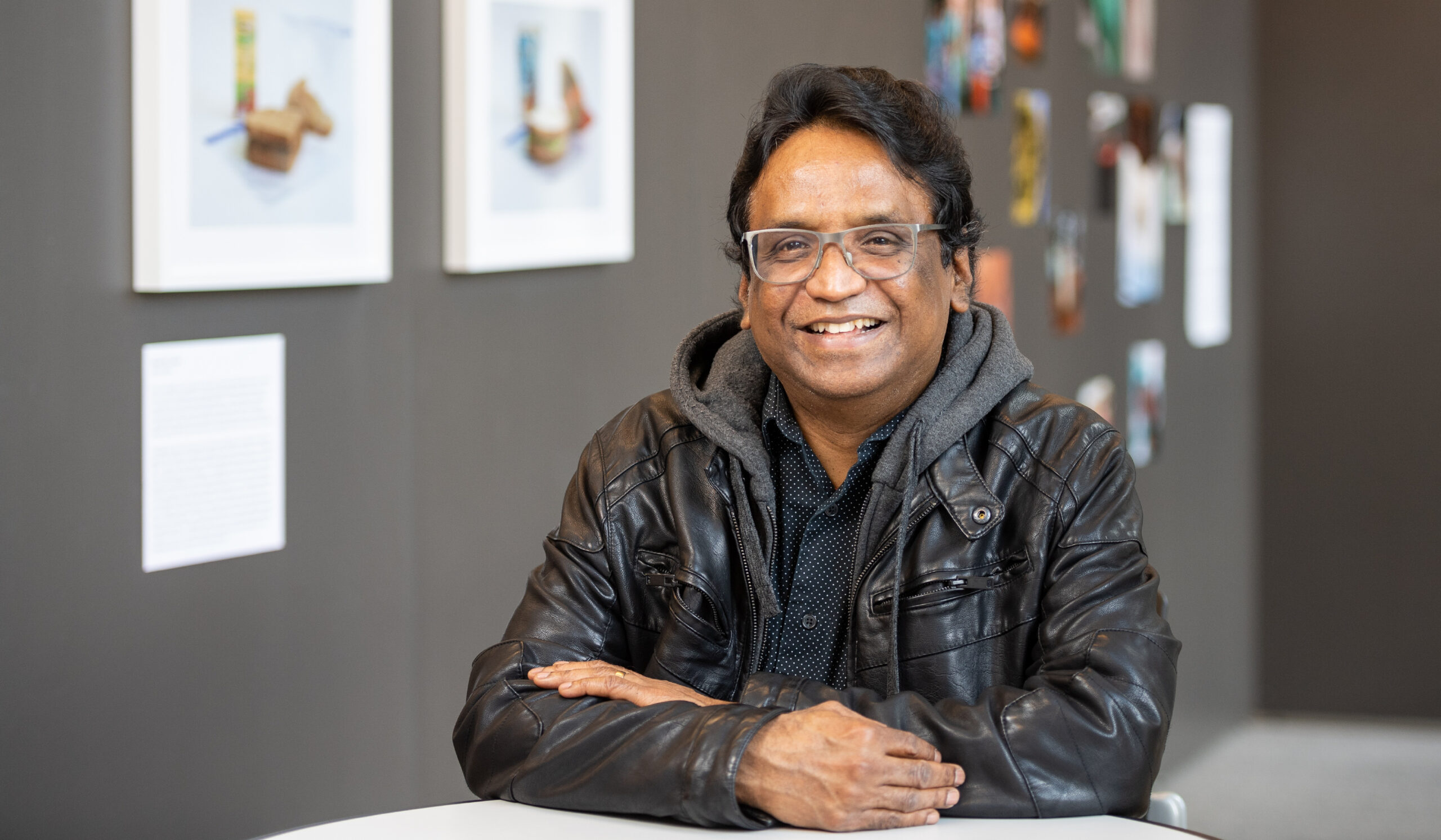 Ramaswami Mahalingam poses at a desk with grey walls in the background. He is wearing glasses and a leather jacket over a sweatshirt and button down.