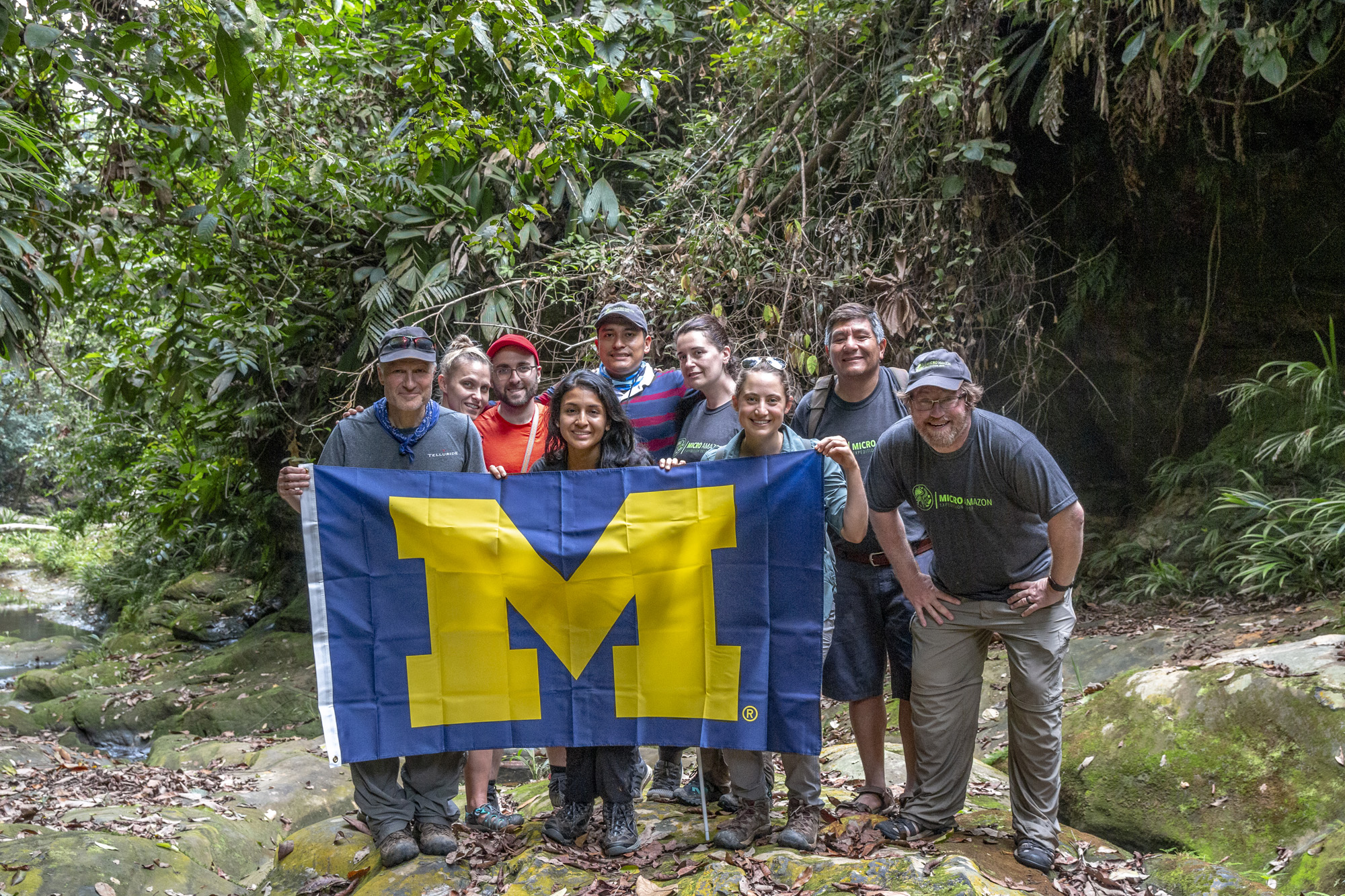 A group of researchers gather behind a University of Michigan flag while on-site in the Peruvian rainforest.