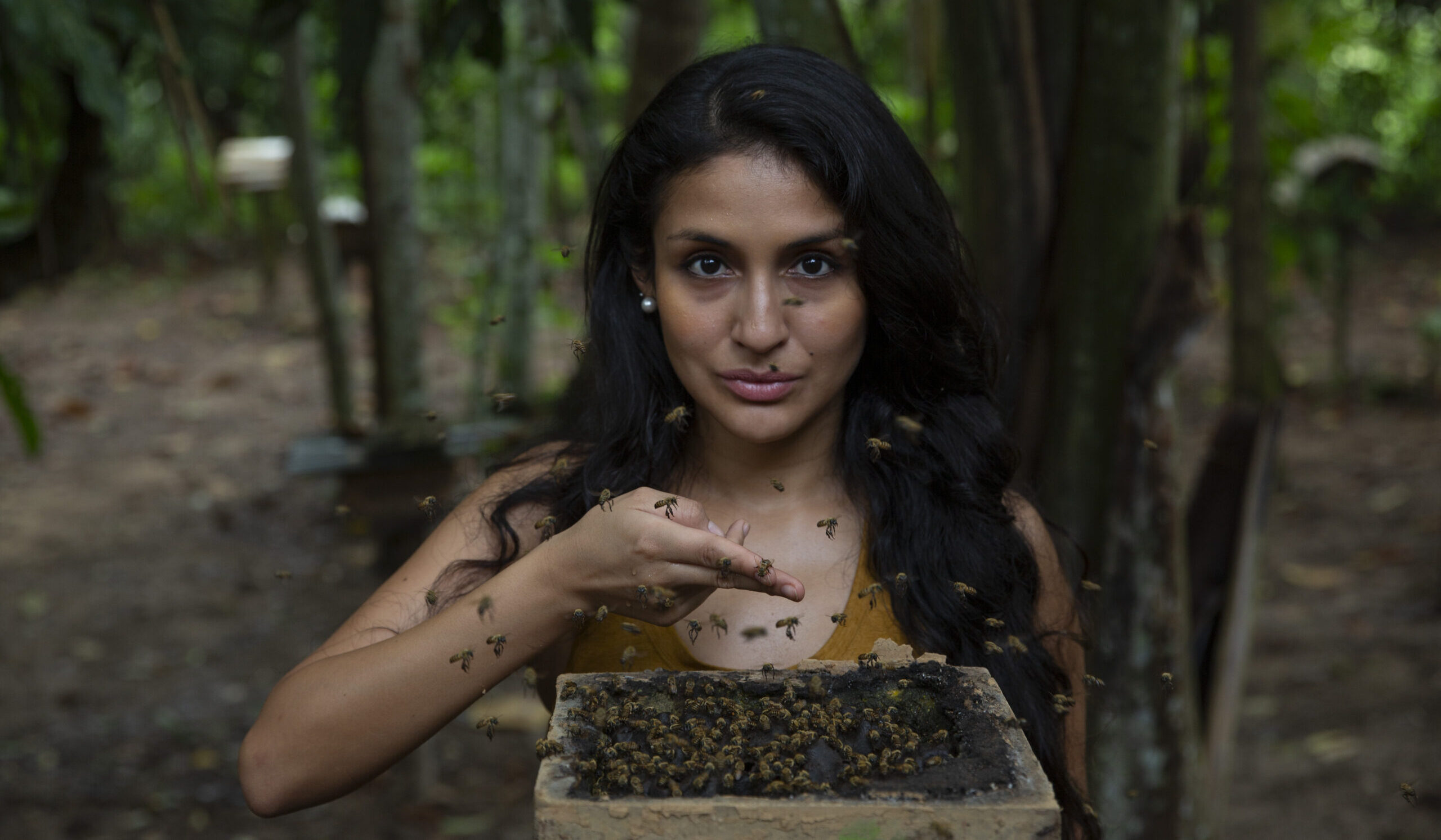 Rosa Vasquez Espinoza holds two fingers out with honey as bees swarm around her in a dark jungle environment.