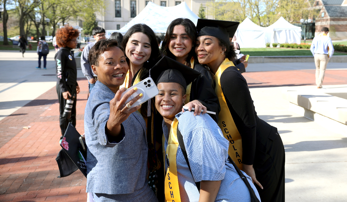 Ayanna (far left) takes a selfie with graduating LEAD Scholars, some of which are wearing graduation caps and stoles, on Ingalls Mall.