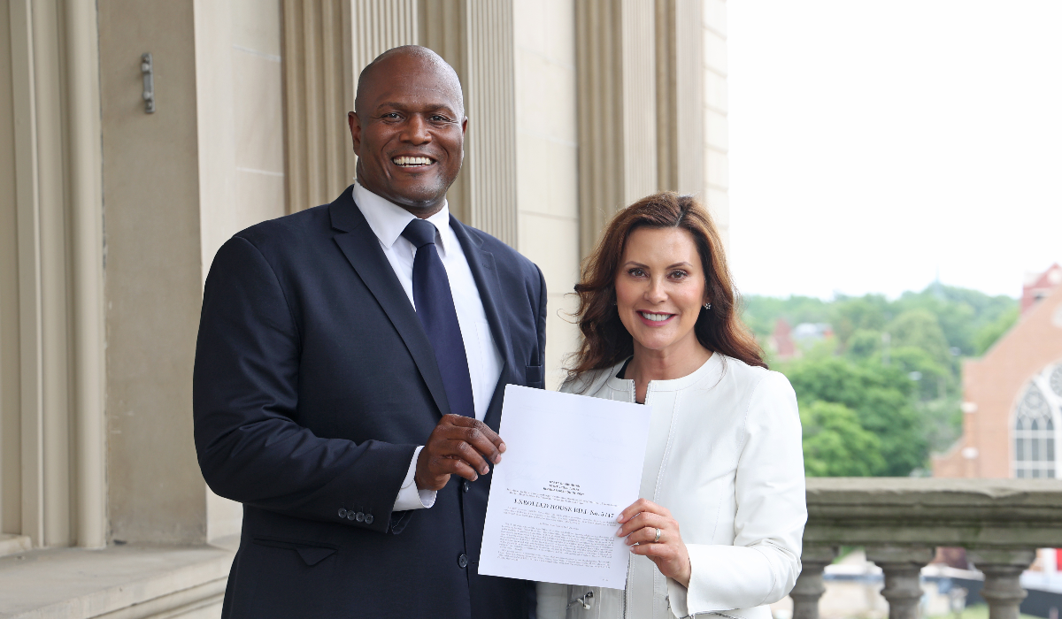 Joe Tate, left, wearing a white shirt and a dark suit, stands with Governer Gretchen Whitmer, who is wearing a white jacket. They are holding a piece of paper that says "Enrolled House Bill No. 5447."