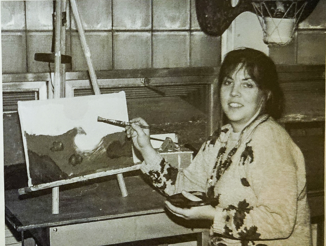 A black and white image of Tammi Browning painting a wave on a canvas on a desk easel.
