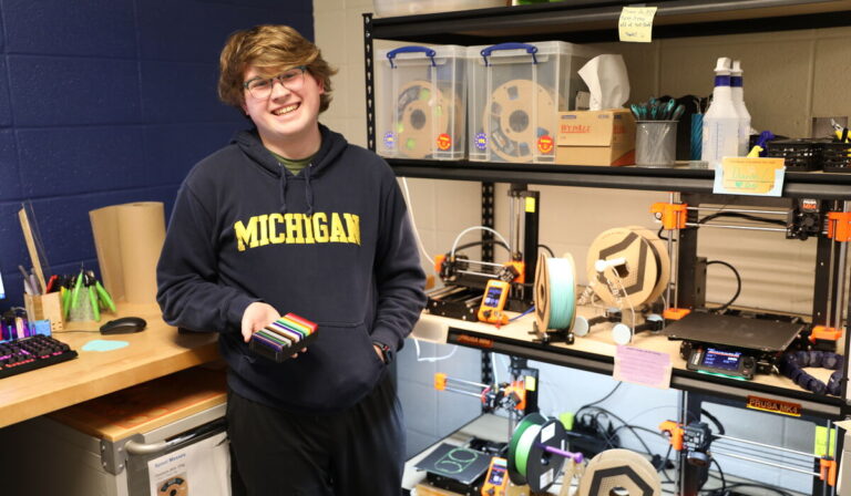 A student wearing a blue Michigan sweatshirt is holding colorful pieces of plastics inside a lab with various items around them.