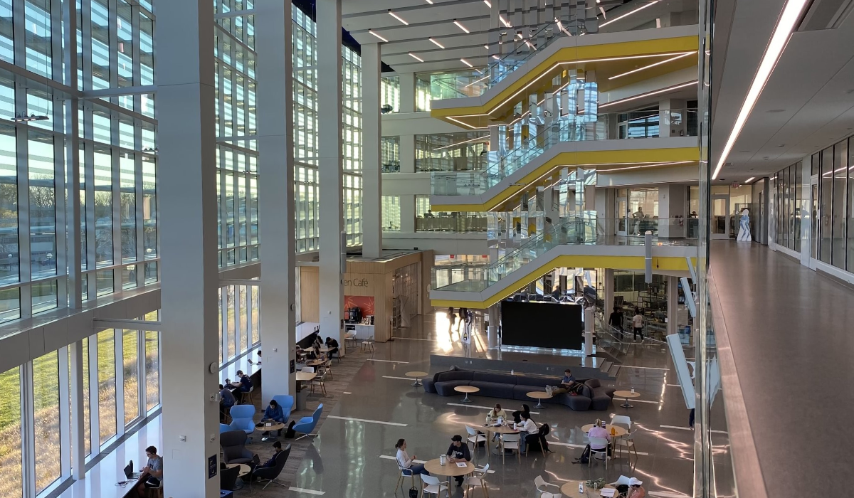 Inside of the Ford Robotics Institute. Taken from the second floor over looking the atrium, there is a tall wall of windows on the left, students gathered at tables on the ground floor, and a winding yellow staircase in the middle.