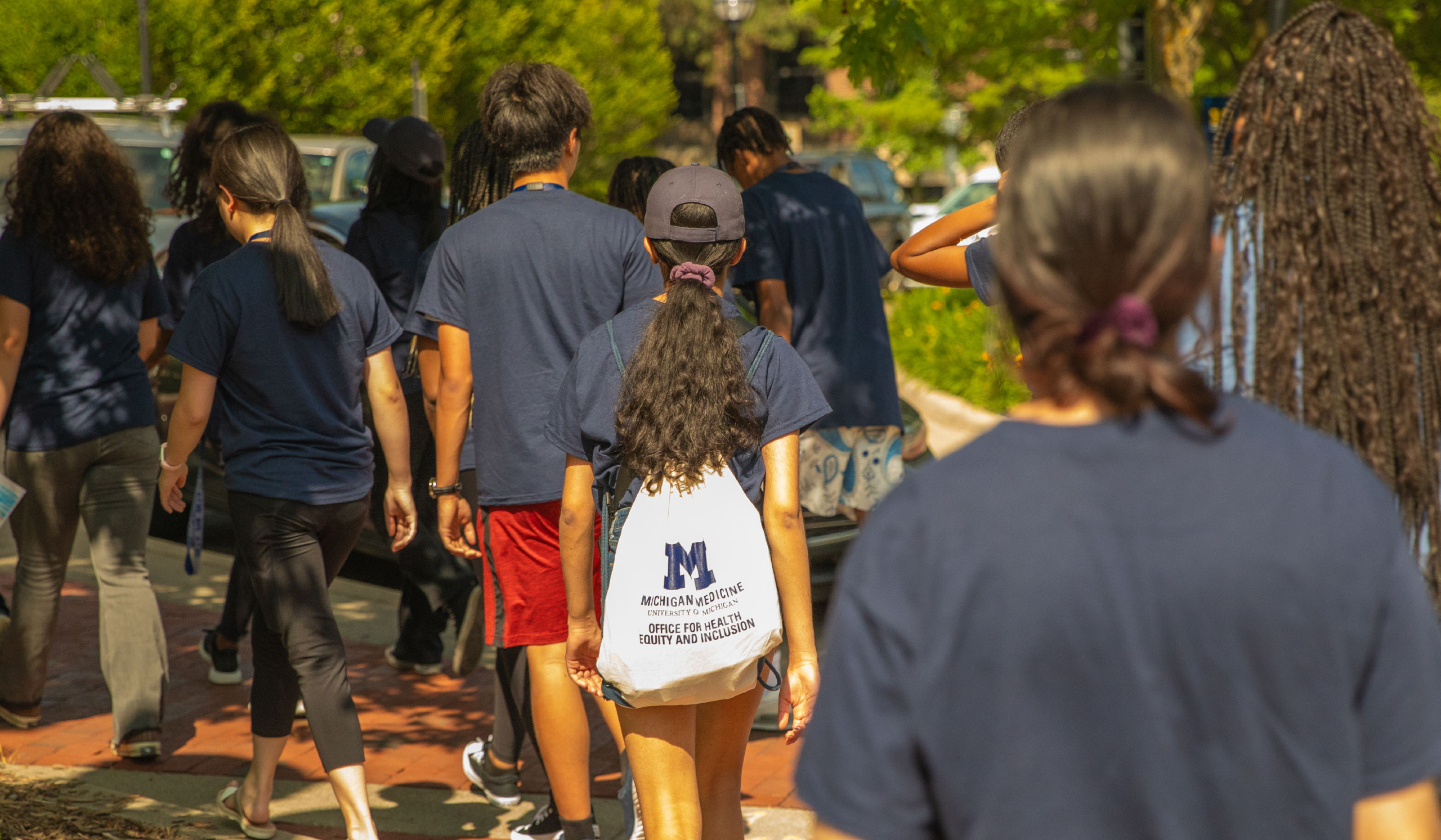 Students in navy shirts walk on a sidewalk at the University of Michigan. In the center, one student has a backpack with a Block M on it.