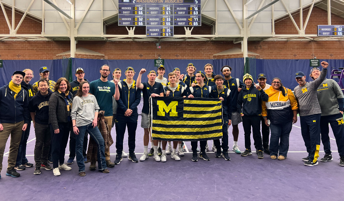 Members of the UM Club of Seattle gather on a tennis court and pose with a striped U-M flag.
