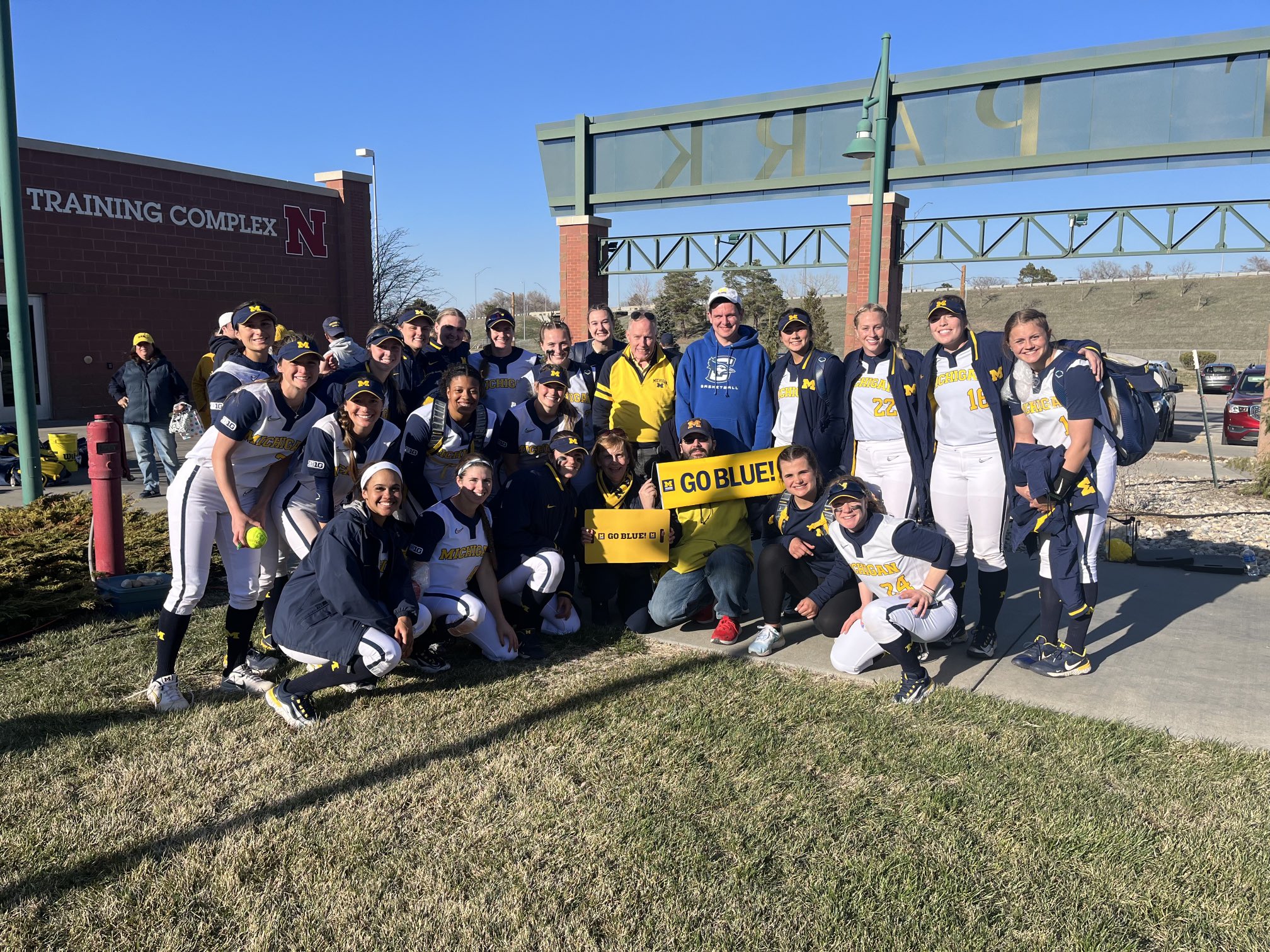 Members of the UM Club Of East Nebraska pose with U-M softball players and "Go Blue" signs outside a brick sports complex.