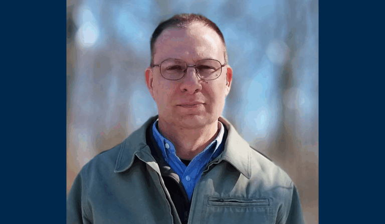 A headshot of the author outdoors with a blurred background. He has short hair and is wearing glasses.