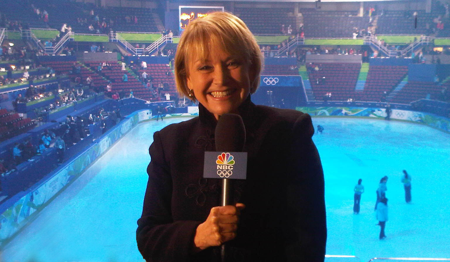 Joyce, in a black jacket, holds a NBC Olympics microphone and poses in front of an ice skating rink from above.