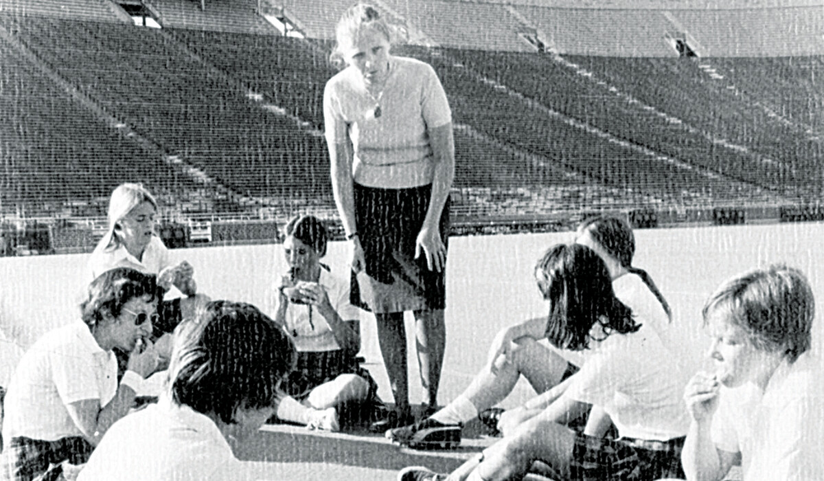 Head Coach Phyllis Weikart (standin) coaches as we are sprawled on the tartan turf of Michigan Stadium during halftime of a home match Fall 1973.