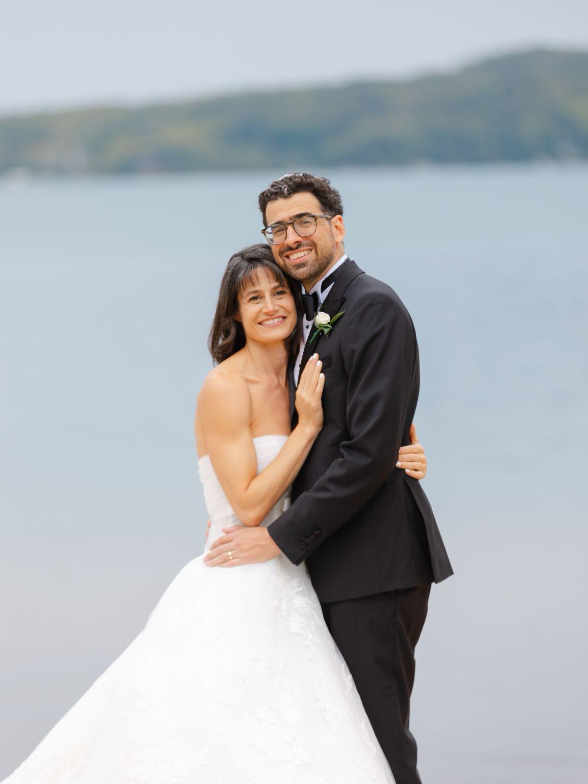 Anne, in a white wedding dress, and Ian, in a black tux, hold each other and pose for a photo with Walloon Lake in the background. 