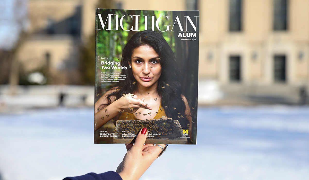 A person holds up a Michigan Alum magazine outside on a snowy day. The cover shows someone in a yellow top, with bees on and around them, standing in front of lush green trees. Prominent headlines about conservation, education, and leadership appear on the cover.
