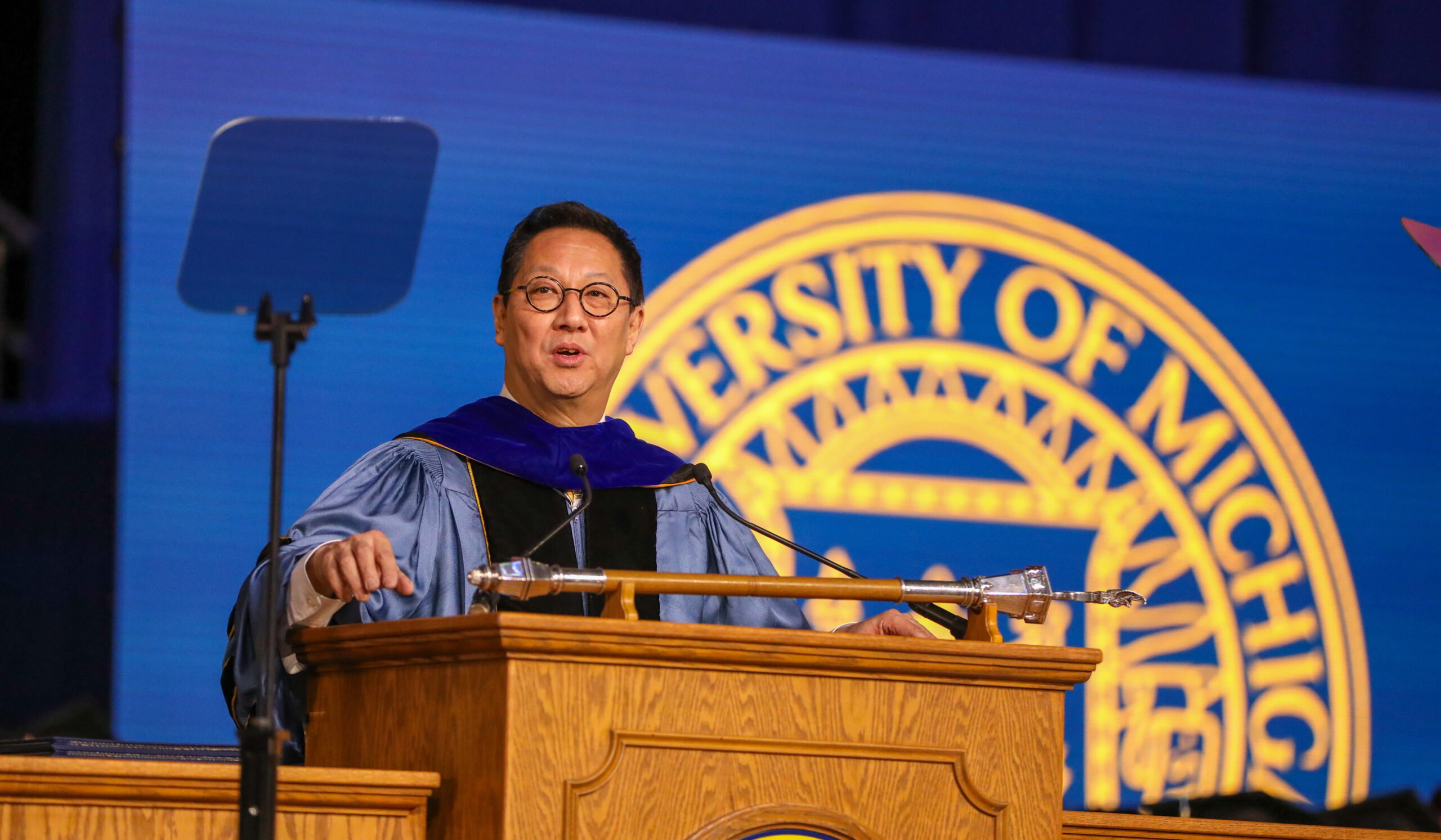 Santa Ono speaks at a brown podium in commencement robes with a large U-M emblem behind him.